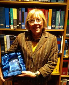 Woman with short blonde hair, wearing glasses and a striped jacket, is holding a box with blue material with a glass etched book in it.
