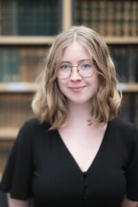Head and shoulders of person with shoulder length blonde hair, glasses, smiling wearing a black top