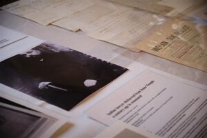 Documents laid out on a table. Text is blurred. One photograph shows a man with glasses and a beard in a suit.