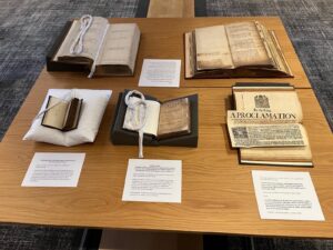 Five different sized books, open, on a wooden table, with labels by each (text unreadable). One book has a piece of paper attached to the page with text: By the King. A Proclamation.