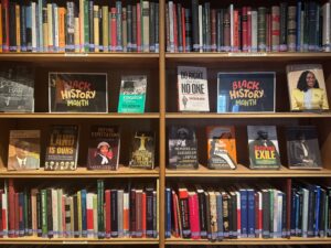Four wooden shelves, top and bottom shelves full or books, spines facing outwards. Middle two shelves with covers of books facing outwards and signs with Black History Month written in red, yellow and green writing.