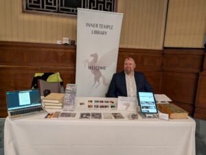 Man sat behind a table with a white table cloth and on it a tablet, a laptop, books, bookmarks and postcards.