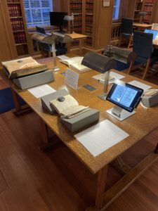 Wooden table with wooden shelves behind it. Open books laid out with a tablet showing the ‘Melissomelos, or the Bee’s Madrigal’.
