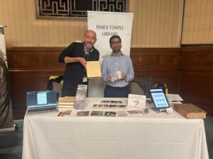 Table with white tablecloth, covered in bookmarks, postcards, books, a laptop and a tablet. Two men stood behind the table, one holding a book the other holding a postcard. Banner behind them reads Inner Temple Library.