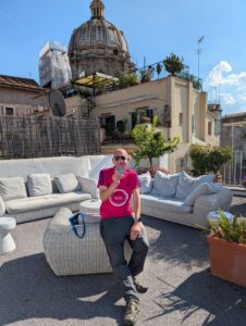 A man sat against a table in a red running top holding a medal, against white outdoor sofas and an old cream building behind