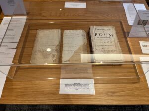 Three pages from a book, Paradise Lost 1688, laying on a wooden table. One page has the title: Paradise Lost A Poem in Twelve Books. The Authour John Milton. The other two pages have handwritten notes including a list of names.