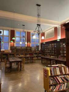 Large room with ochre walls, large windows and wooden bookshelves in the background, and a trolley full of books in the foreground. Metal light fixings hang from the ceiling.