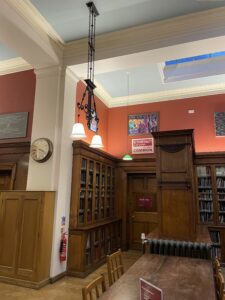 Ochre walls, white celing, dark wooden book shelves, doors and table. Pictures hang on the walls, metal light fixings hang from the ceiling