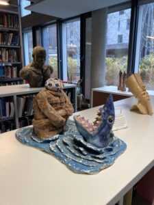 Ceramic of a shark's head, mouth wide open with teeth showing opposite a person in a straightjacket with what looks like a small head made up of eyes.
