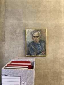 Painting of a man, elbows up, hanging on a wal. with a box file of folders in front of him. 