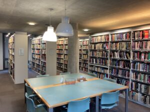 Room full of grey shelving full of books. Light blue table in front with light blue chairs and light grey lights hanging from the concrete ceiling