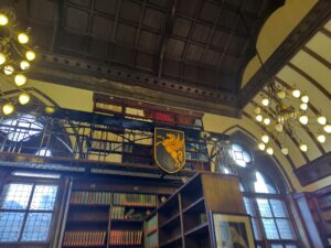 Dark wooden ceiling, a chandelier light hanging down, books shelves on a gallery above and bookshelves below. A Pegasus in a shield attached to the iron gallery.