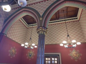 Wall painted one half in red with the Inn's shields painted on, the top half with cream and lines of red with a leaf or flower pattern repeated. In front is an archway with gold details and painted with reds and greens in a geometric pattern