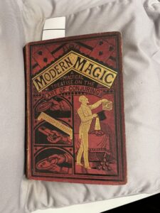 Book cover, red with pictures of playing cards, hands holding various instruments of magic and a gold figure holding a bird cage and a top hat. Text: Modern Magic, A Practical Treatise on the Art of Conjuring