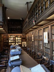 Room with dark wooden shelving filled with books and a long table with wooden chairs in the centre. Open books are on display on the table and in a glass cabinet at one end of the room.